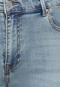 Lys blå denimjeans i nærbilde som viser frontlomme, beltehempe, metallknapp og detaljert søm.