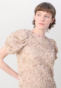 Beige Spitzenoberteil mit Puffärmeln, das mit komplexen Blumenmustern und einer strukturierten Oberfläche versehen ist, einen runden Ausschnitt und transparente Details aufweist.