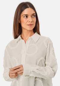 White blouse with textured, ruffled circular patterns, button front, long sleeves, and a classic collar. Lightweight fabric with a soft finish.