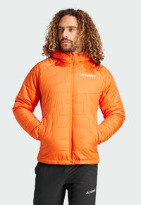 Oranžová zateplená bunda se zipem, kapucí a logem. Má prošívaný design a dvě boční kapsy. Nositá přes černé kalhoty.