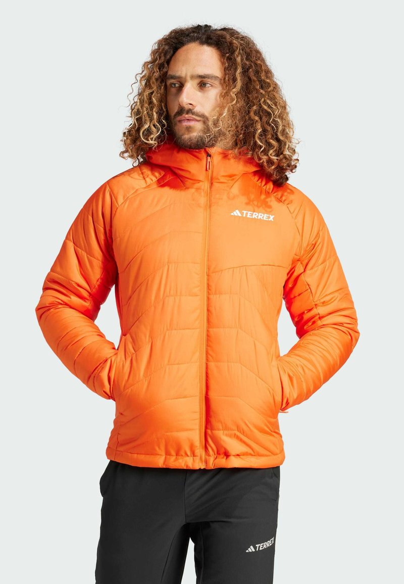 Oranžová zateplená bunda se zipem, kapucí a logem. Má prošívaný design a dvě boční kapsy. Nositá přes černé kalhoty.