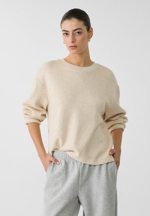 Pull en tricot beige surdimensionné avec un col rond côtelé et des poignets roulés, associé à un pantalon de sweat gris clair, mettant en valeur une texture douce.