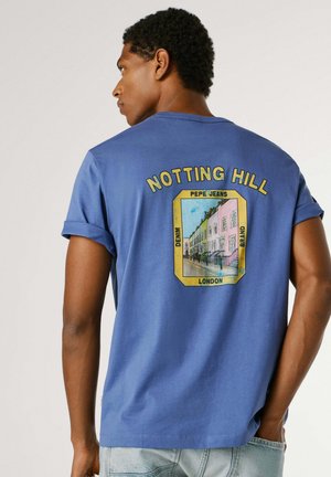 Mann trägt blaues T-Shirt mit hochgekrempelten Ärmeln, das auf der Rückseite "Notting Hill" und eine farbenfrohe Grafik einer Londoner Straße zeigt.