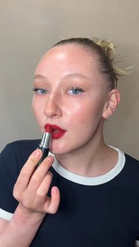 Personne à la peau claire et aux yeux bleus appliquant du rouge à lèvres rouge, portant une chemise noire avec un col blanc, sur un fond beige uni.
