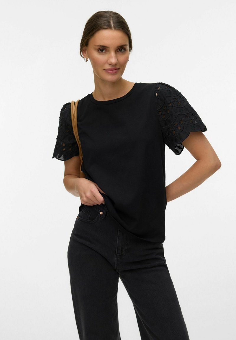 Kobieta ubrana w czarną bluzkę z koronkowymi rękawami o falistym brzegu oraz czarne dżinsy, stojąca z ręką w kieszeni, widoczny jasnobrązowy pasek torby na ramię.
