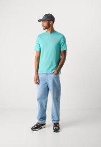Mand i teal grafisk t-shirt, lyseblå jeans, multicolor sneakers og mørk kasket, stående med den ene hånd i lommen og kiggende til venstre på hvid baggrund.