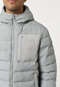 Svetlošedý puffa kabát s kapucňou, predným zapínaním na zips a vreckom na hrudi. Vystužená textúra so šitými švami.