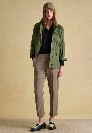 Jauna moteris dėvi smėlio spalvos kepurę, žalią striukę, juodą marškinėlį, languotas kelnes ir juodai baltus loaferius, stovinčią priešais neutralią fono sieną.