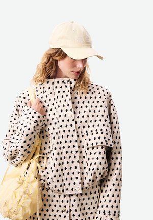 Femme portant une casquette beige et une veste crème à pois noirs irréguliers, tenant un sac à cordon jaune pâle sur l'épaule.