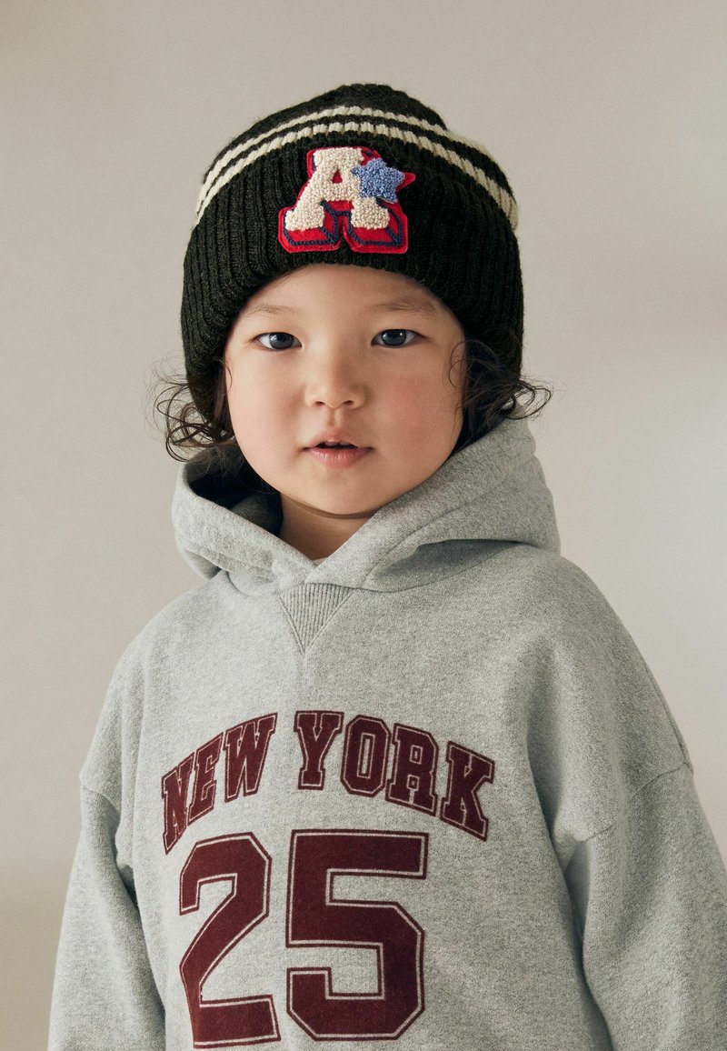 Dunkelgrüne Strickmütze mit weißen Streifen und einem strukturierten roten und beigen "A"-Patch; grauer Kapuzenpullover mit "NEW YORK 25" in maroon.