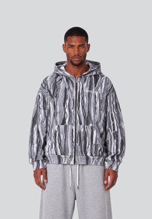 Mann trägt eine grau-schwarz gemusterte Hoodie mit Reißverschluss und Kapuze sowie hellgraue Jogginghose, steht vor einem schlichten Hintergrund.