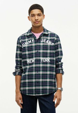 Giovane uomo che indossa una camicia a quadri verde e bianca con bottoni con testo "Tommy Jeans New York" e un orologio da polso argentato.