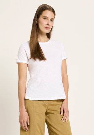 Frau mit langen braunen Haaren, die ein weißes T-Shirt mit kurzen Ärmeln und hellbraune Hosen trägt, steht vor einem beigen Hintergrund.