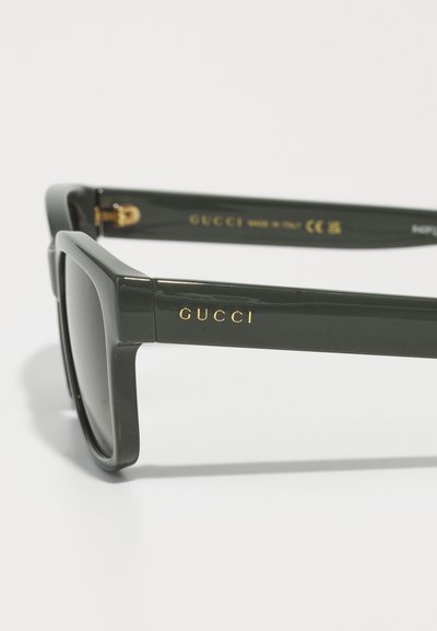 Ochelari de soare verzi cu ramă pătrată, finisaj lucios, logo auriu "GUCCI" pe bratul ochelarilor și lentile întunecate. Fabricat în Italia.