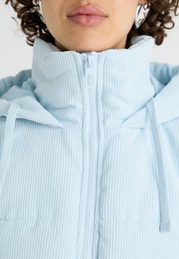 Helleblaue Cordjacke mit hohem Kragen, Frontreißverschluss und Kordelzugakzenten, die durchgehend eine vertikale Rippstruktur aufweist.