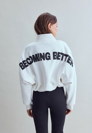 Kvinde iført en hvid sweatshirt med "BECOMING BETTER" i sorte bogstaver på ryggen og sorte tætsiddende bukser, vendt væk fra kameraet.