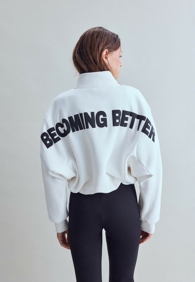 Femme portant un sweat blanc avec "BECOMING BETTER" en lettres noires dans le dos et un pantalon noir ajusté, tourné dos à la caméra.