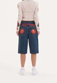 Shorts de mezclilla azul con grandes gráficos de manzana roja en los bolsillos, con un logo bordado de "Ed Hardy", dobladillo enrollado y corte relajado.