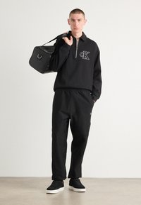 Crna zip-up dukserica s "CK" logotipom na prsima, uparen s crnim suženim trenirkama. Crna torba s ravnim dnom, držana u desnoj ruci, i crne cipele.