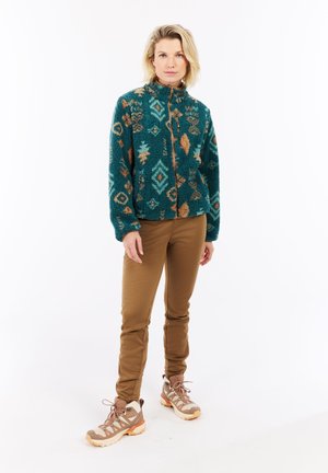 Vrouw die staat in een teal-gepatenteerde fleecejack, bruine broek en beige wandelschoenen op een witte achtergrond.