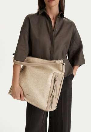 Donna con maglietta e pantaloni scuri tiene una grande borsa beige con tasca con cerniera frontale e tracolla.