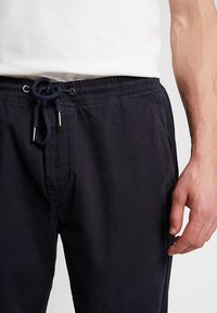 Meripuku, jossa on elastinen vyötärö, säädettävä nyöri ja sivutaskut. Kangas on sileä ja rento muotoilu.