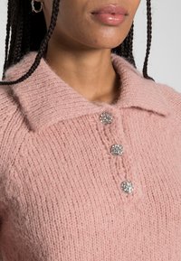 Ljusrosa stickad tröja med krage, prydd med tre silverknappar. Mjuk textur och en bekväm, avslappnad passform.