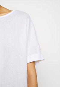 Valkoista pellavapaitaa, jossa on pyöreä kaula-aukko ja lyhyet, käännetyt hihat. Kankaan pinnassa on hieman tekstuuria, mikä luo rennon ilmeen.