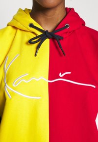 Personne portant un sweat à capuche bicolore avec le côté gauche jaune et le côté droit rouge, des cordons noirs et une broderie blanche en cursive sur la poitrine.