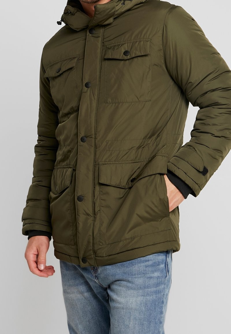 Olivgrön vadderad jacka med huva, knäppning fram, två bröstfickor och elastiska ärmslut. Strukturerad tyg med en slät yta.