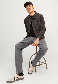 Gray denim jacket over a black t-shirt, paired with gray jeans and beige sneakers. The model sits on a stool, wearing a black cap.