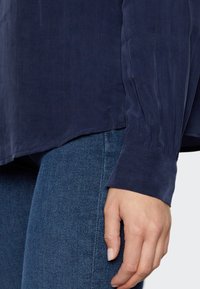 Granatowa bluzka z długim rękawem, wykonana z lekkiego materiału, charakteryzująca się luźnym krojem, plisowanymi mankietami i zaokrąglonym dołem, w zestawie z niebieskimi dżinsami.