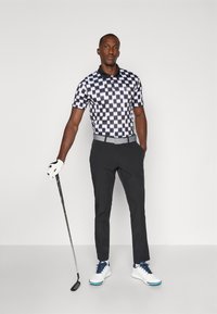 Polo noir et blanc à carreaux associé à un pantalon noir et des chaussures de golf blanches. Comprend un club de golf à la main et un gant blanc.