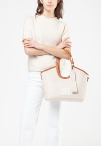 Borsa tote in pelle beige con accenti marroni, forma strutturata, chiusura con zip e un'etichetta con logo. Il modello tiene la borsa indossando un maglione chiaro.