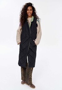 Schwarze gesteppte lange Weste mit grünem Kragen, ausgestattet mit einem Frontreißverschluss und seitlichen Taschen, kombiniert mit einer Hose im Leopardenmuster und braunen Schuhen.
