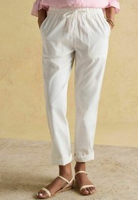 Pantaloni albi din bumbac cu talie elastică și șnur, prevăzuți cu două buzunare frontale și tivuri suflecate. Purtați cu sandale bej.