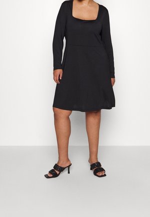 Vrouw draagt een zwarte jurk tot op de knie met lange mouwen en een vierkante halslijn, gecombineerd met zwarte sandalen met hak en ruches aan de banden.