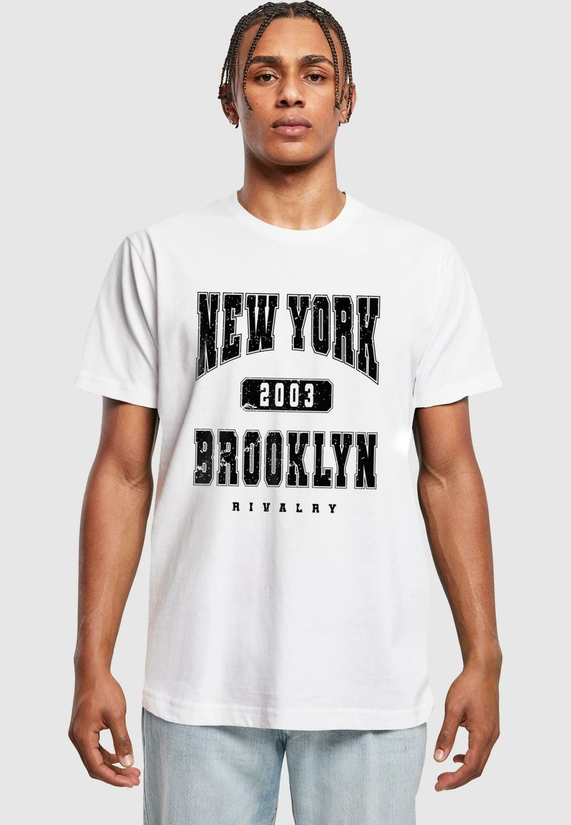 Balta medvilninė marškinėlių su drąsiais juodais užrašais: "NEW YORK," "2003," "BROOKLYN," ir "RIVALRY." Trumpi rankovių ir laisvas siluetas.