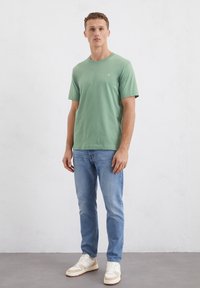 T-shirt in cotone verde con colletto rotondo, maniche corte e un piccolo logo. Abbinato a jeans azzurri e sneakers bianche con dettagli beige.