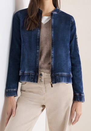 Femme portant une veste bomber en denim bleu délavé sur un pull col V taupe et un pantalon beige taille haute, debout contre un mur clair.