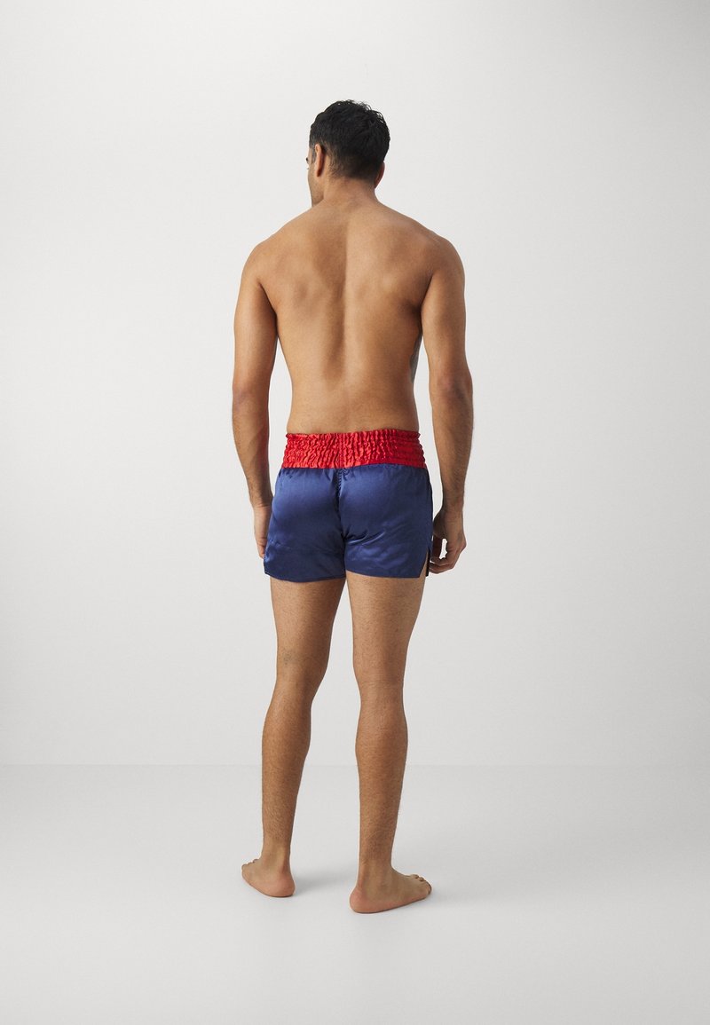 Homme torse nu, debout pieds nus, de dos, portant un short bleu avec une large ceinture élastique rouge, sur un fond blanc uni.