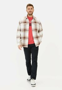 Rutig flannel-skjorte i hvid, rød og brun over en rød grafisk t-shirt, parret med sorte bukser og hvide sneakers, stående mod en ensfarvet baggrund.