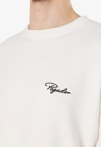 Hvid struktureret crew-neck sweatshirt med et sort broderet "Pegador" logo på brystet.
