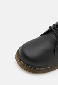 Scarpa in pelle nera con punta arrotondata, texture e dettagli in cucitura gialla. Presenta una suola a rilievo per la trazione e un robusto design con lacci.