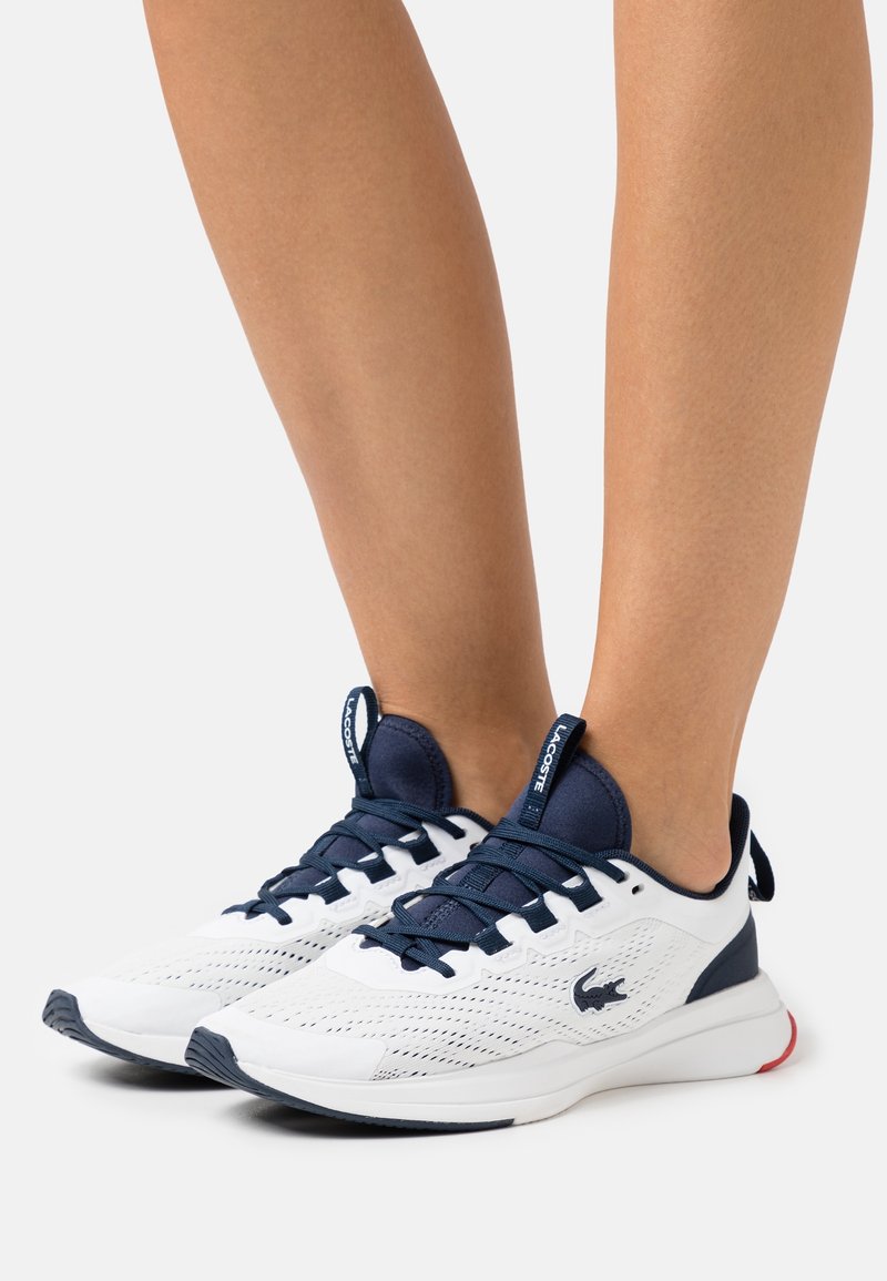 Chaussures de sport blanches avec des accents bleu marine, dessus en mesh, semelle intermédiaire texturée et détails rouges au talon. Dotées de lacets et d'un logo de marque sur le côté.