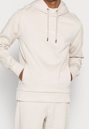 Mann trägt hellbeigen Calvin Klein Hoodie und passende Jogginghose, die Hände in den Taschen des Hoodies vor einfachem Hintergrund.