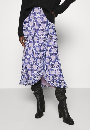 Personne portant une jupe midi fleurie violette et blanche, des bottes noires à talons hauts décontractées et un pull noir, une main sur la hanche.