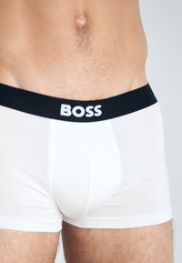 Weiße Boxer-Briefs mit einem schwarzen Bund, auf dem der Markenname "BOSS" in fetten weißen Buchstaben steht. Aus weichem, dehnbarem Material gefertigt.