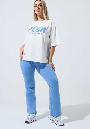 Kobieta ubrana w białą, oversize'ową koszulkę z niebieskim napisem, jasnoniebieskie spodnie i białe sneakersy, stojąca z rękami na biodrach na białym tle.