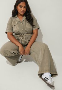 Tans färgad utility jumpsuit med korta ärmar, insvängd midja, två framfickor och vida ben. Kombinerad med svart och vit sneakers.